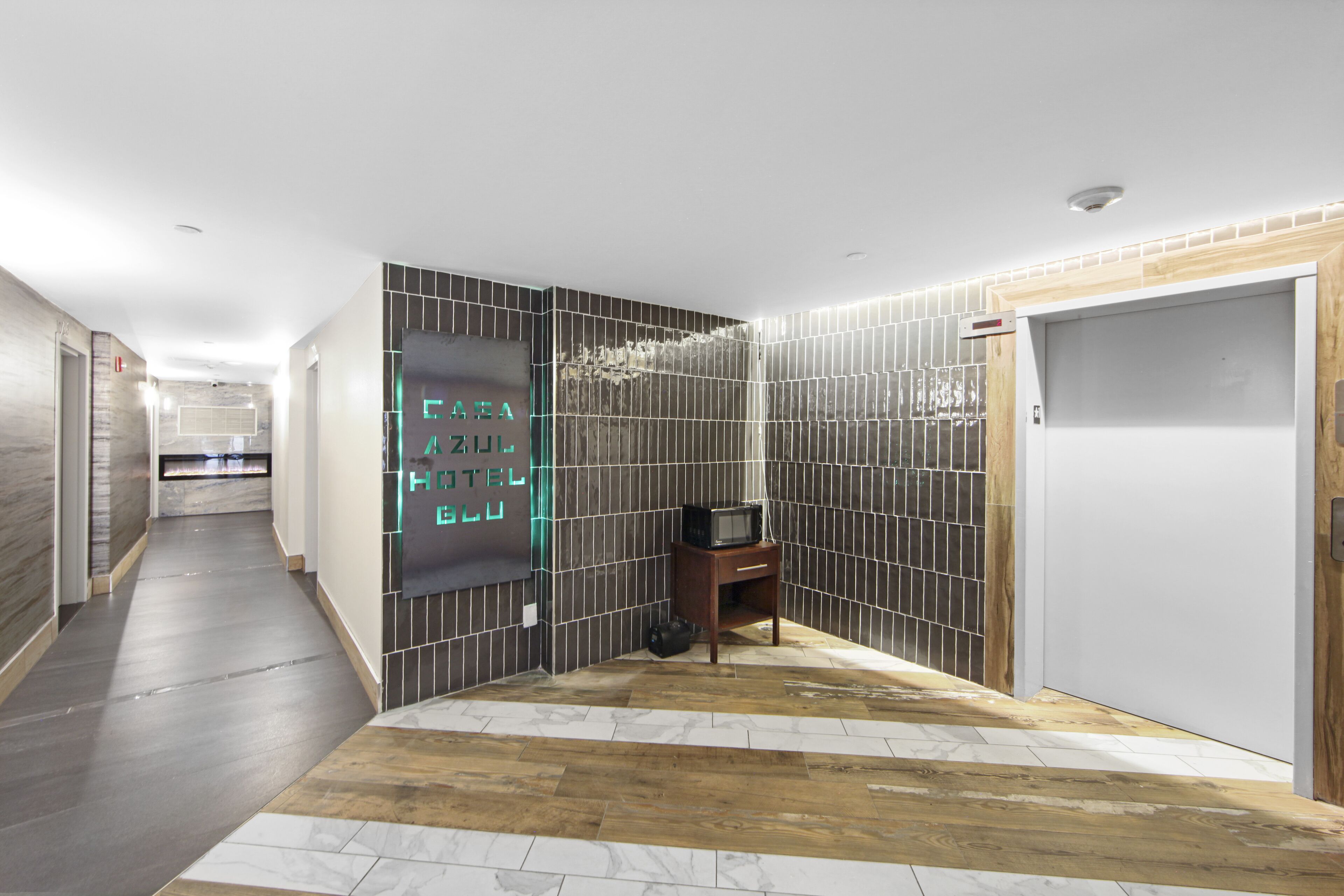 A hotel hallway with a modern design, featuring a wooden floor, tiled walls, and a neon sign.