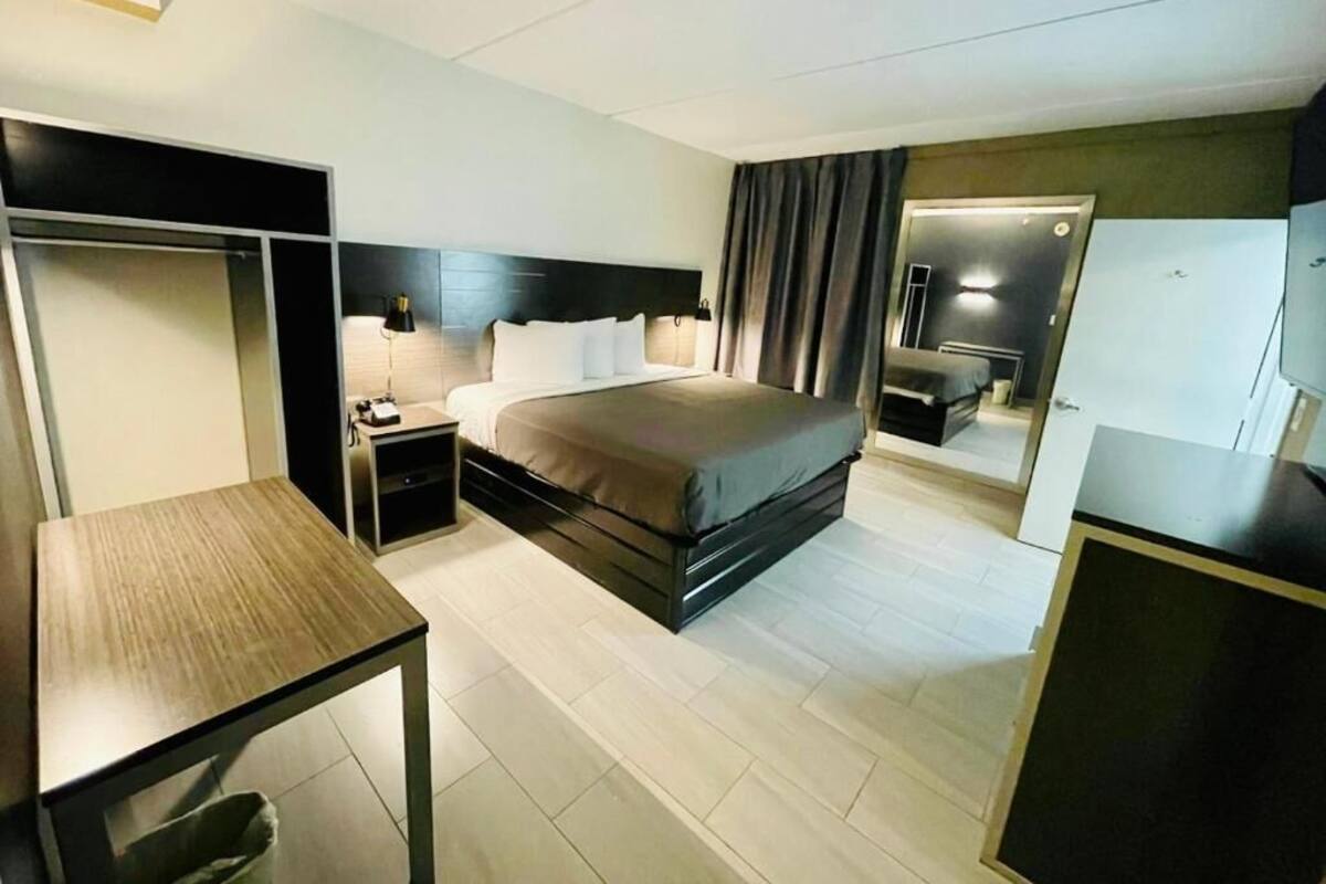 A modern hotel room with a bed, bedside table, lamp, wardrobe, and a mirror.