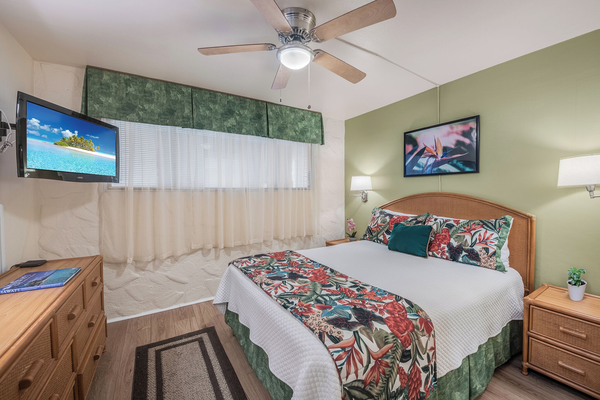 Un dormitorio con cama, ventilador de techo, televisor, mesita de noche, cómoda y ventana con persianas.