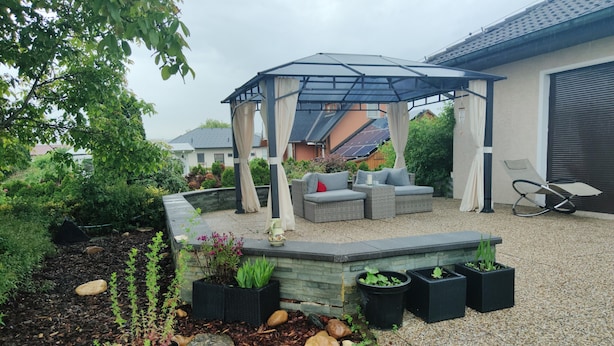 Ein überdachter Außenbereich mit Pergola, einer Couch und einem Lounge-Sessel.