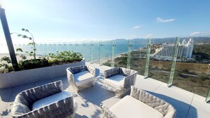 A balcony with two cushioned chairs and a small table, overlooking a coastal view with a glass railing and a distant building.