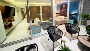 A modern living room with a sofa, a coffee table, and a TV. There is a sliding glass door leading to a bedroom with a bed and a nightstand.