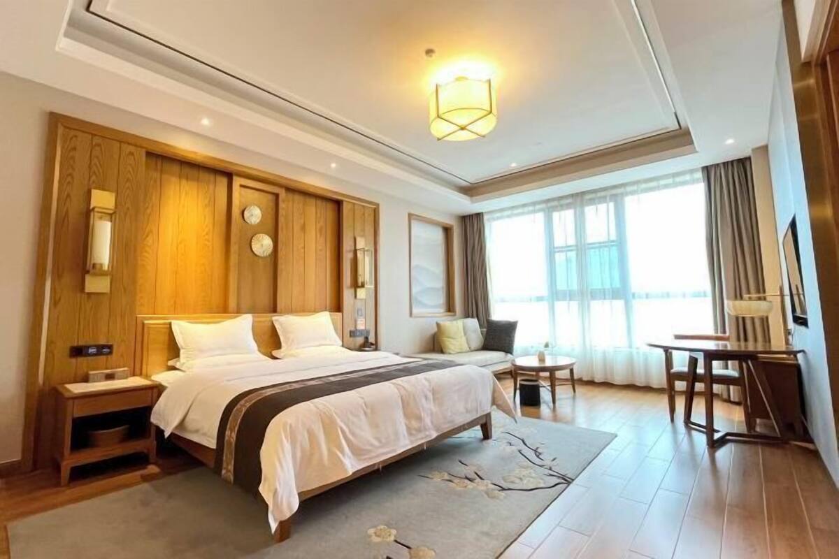 Habitación de hotel con una cama grande, una zona de estar con sofá y sillones, una mesita y un ventanal con cortinas.