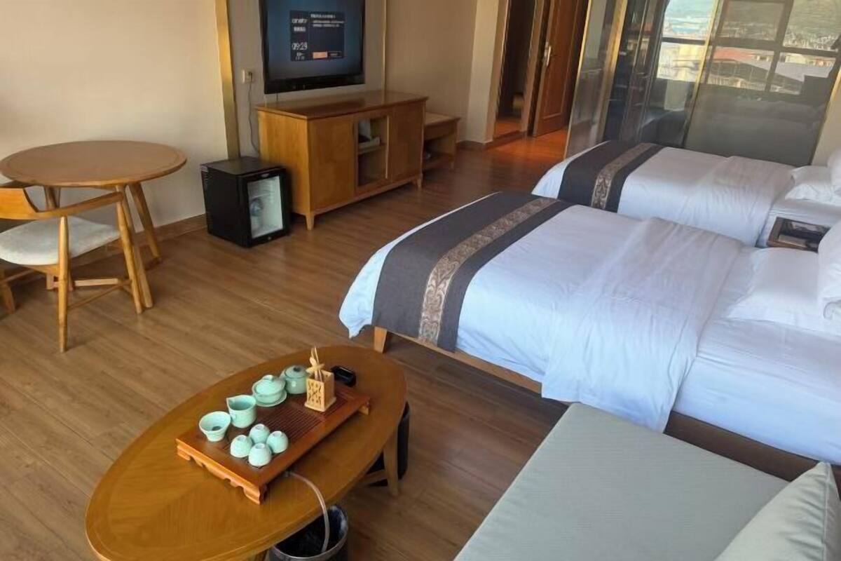 Habitación de hotel con cama, televisor, mesa de madera con una bandeja de tazas de té y vistas a través de un ventanal.