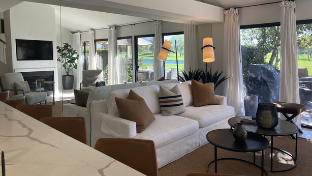 Ein modernes Wohnzimmer mit einem weißen Sofa, einem Flachbildfernseher und einem Kamin. Es gibt einen Essbereich mit Tisch und Stühlen sowie große Fenster mit Blick ins Freie.