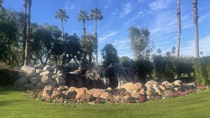 Ein angelegter Garten mit einem Wasserfall, Palmen und einer Rasenfläche mit rosa Blumen.