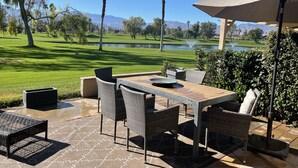 Eine Terrassensitzgelegenheit im Freien mit Korbmöbeln, einem Tisch und einem großen Sonnenschirm mit Blick auf einen Golfplatz und Berge.