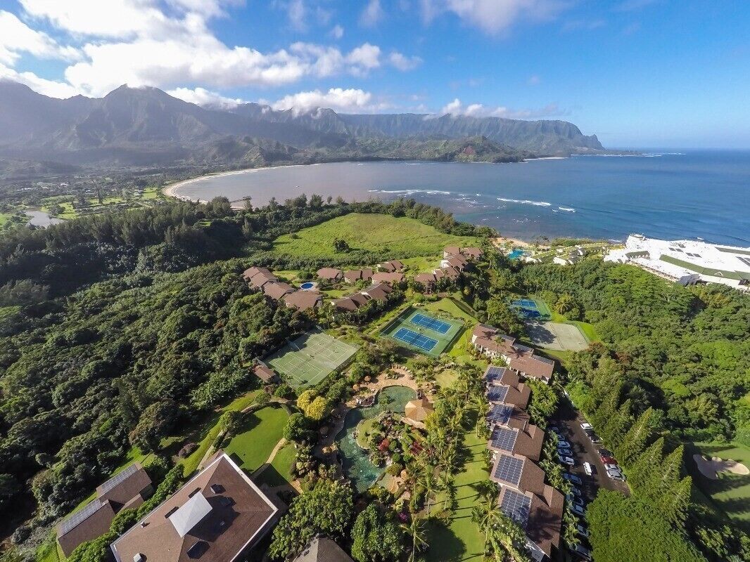 An aerial view of a resort with multiple buildings, a large pool, and a view of the ocean and mountains.