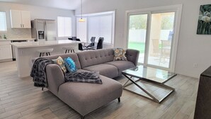 Un salon moderne avec un canapé d’angle gris, une table basse en verre et un coin cuisine doté de meubles blancs et d’un réfrigérateur.