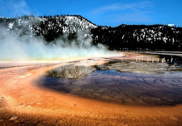 Un'area geotermica con una pozza d'acqua che riflette il paesaggio circostante, vapore che sale dal terreno e una montagna boscosa sullo sfondo.