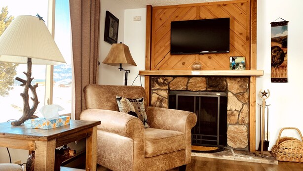 Ein gemütliches Wohnzimmer mit Kamin aus Stein, einem bequemen Sessel mit Zierkissen, einem Holztisch mit Lampe und einem flachbildfernseher, der über dem Kamin angebracht ist.