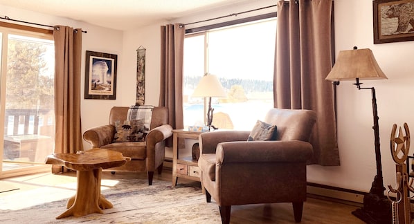 A cozy living room with a sofa, armchair, and a small table. There is a lamp, a picture frame, and a window with curtains.