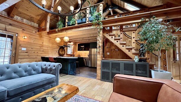 A cozy cabin interior with a living area featuring a tufted sofa and a coffee table, a kitchen with modern appliances, and a wooden staircase. - Modern Rustic Log Chalet w/ mtn views, pool & bonus loft on 17 secluded acres  (Grand Gorge)