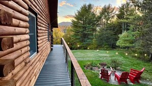 A log cabin with a deck leading to a grassy yard with red Adirondack chairs. - Modern Rustic Log Chalet w/ mtn views, pool & bonus loft on 17 secluded acres  (Grand Gorge)