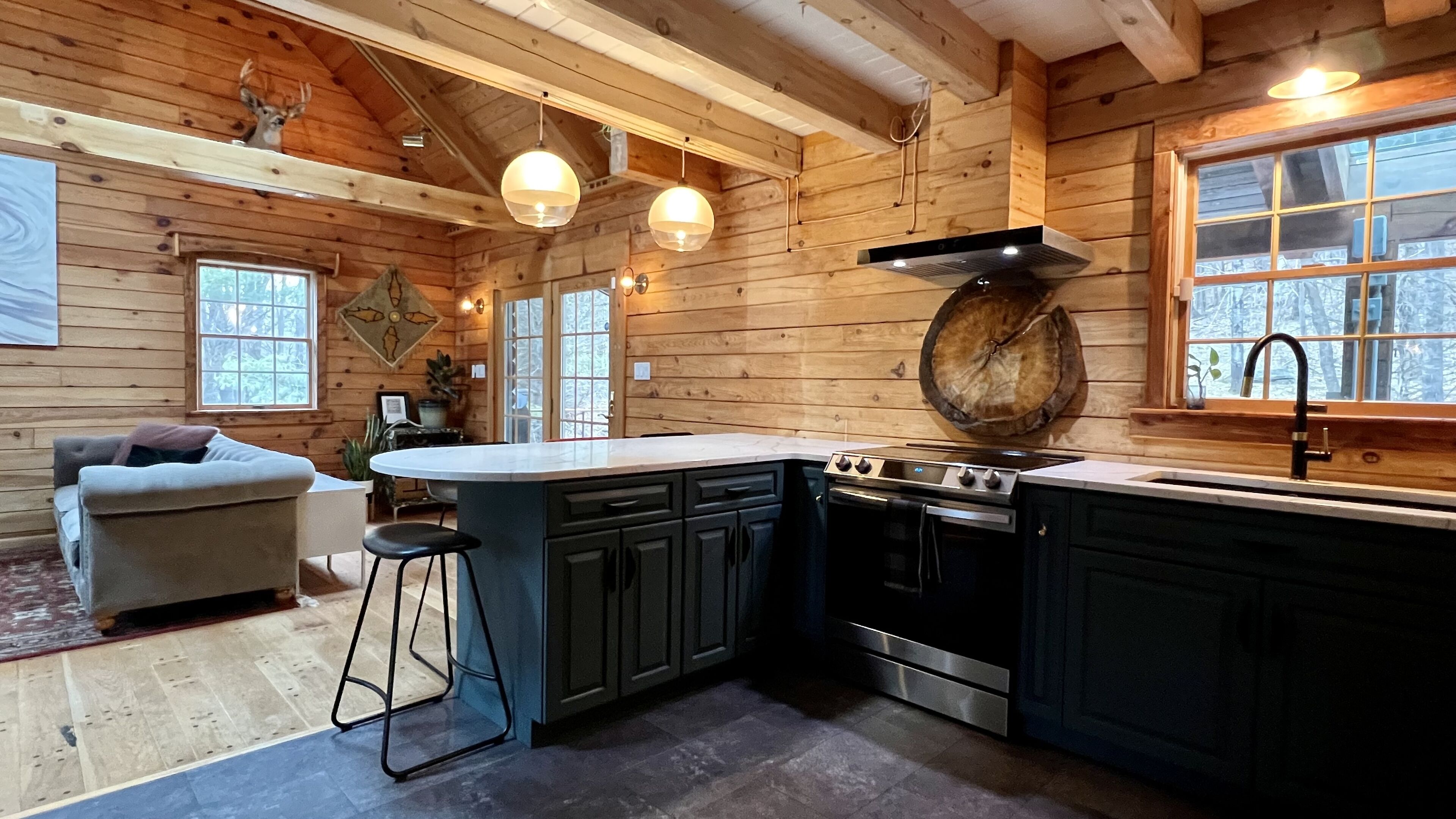 A cozy cabin-style kitchen with wooden walls, a central island, and a dining area with a sofa and bar stool. - Modern Rustic Log Chalet w/ mtn views, pool & bonus loft on 17 secluded acres  (Grand Gorge)