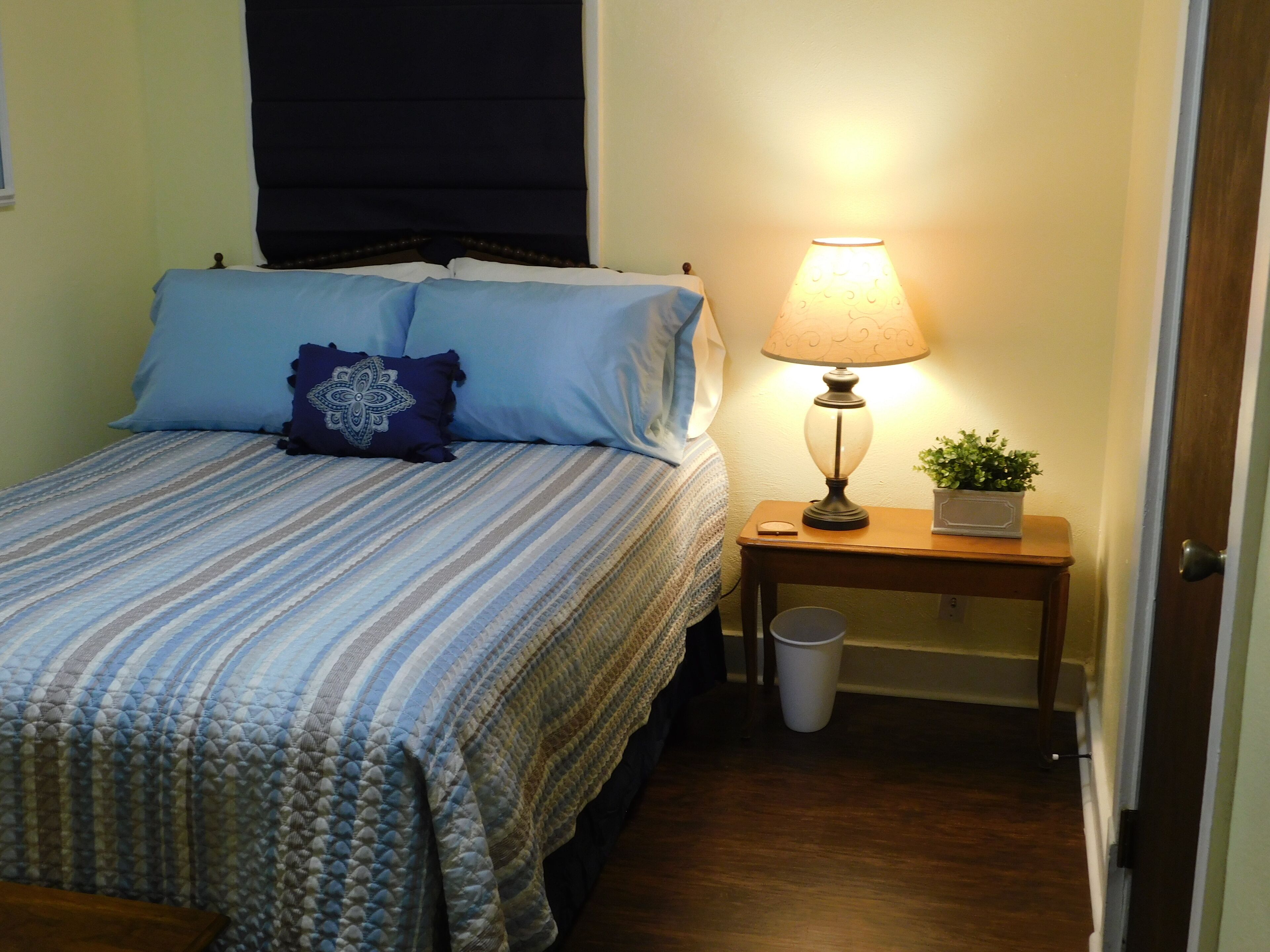 A neatly made bed with a striped bedspread and blue pillows, a bedside lamp, a small table with a plant, and a waste bin.