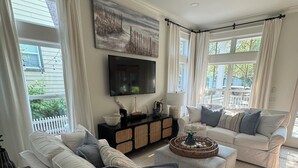 A living room with a large sectional sofa, a smaller sofa, a coffee table, and a flat-screen TV mounted on the wall. There is a window with curtains, a ceiling fan, and a decorative wall piece above the TV.