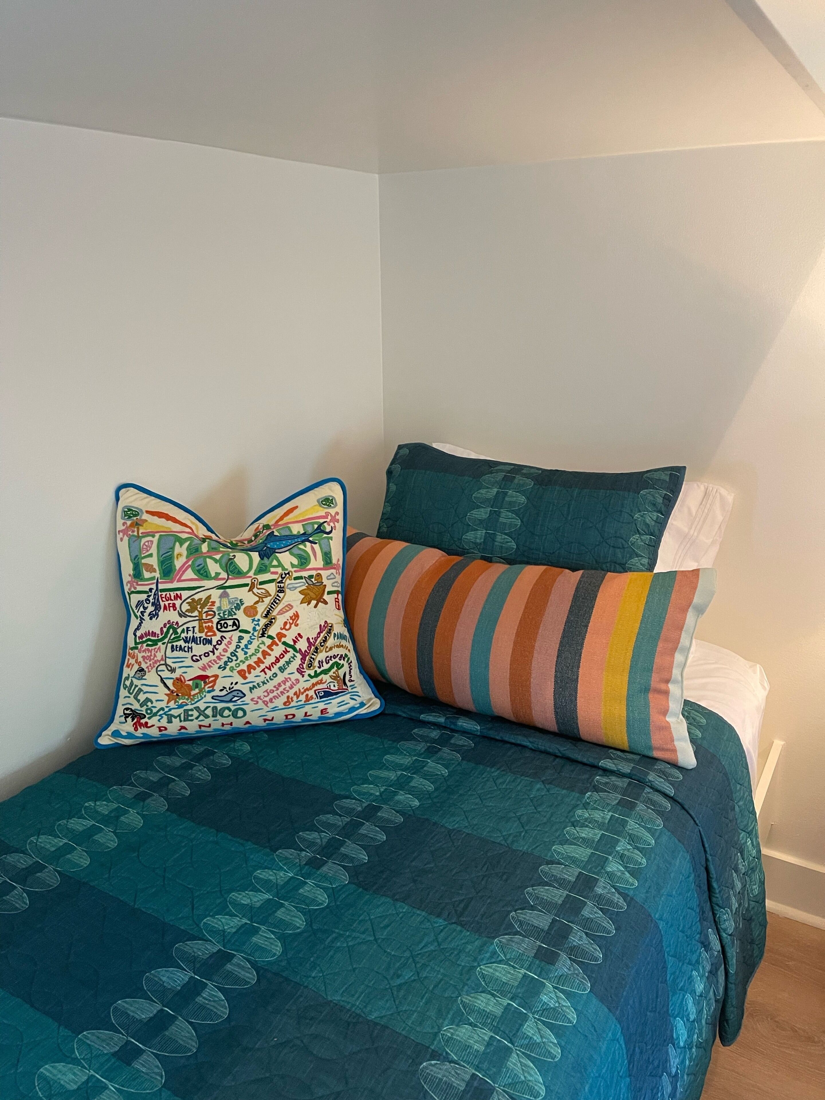 Una cama con un edredón azul turquesa y dos almohadas, una con un diseño de mapa colorido y la otra a rayas.