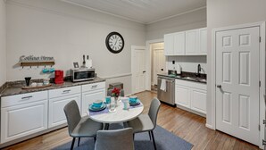 A compact kitchen with white cabinetry, a microwave, and a sink. There is a dining table with chairs and a clock on the wall.