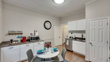 Une cuisine compacte avec des meubles blancs, un four à micro-ondes et un évier. On y trouve une table à manger avec des chaises et une horloge au mur.
