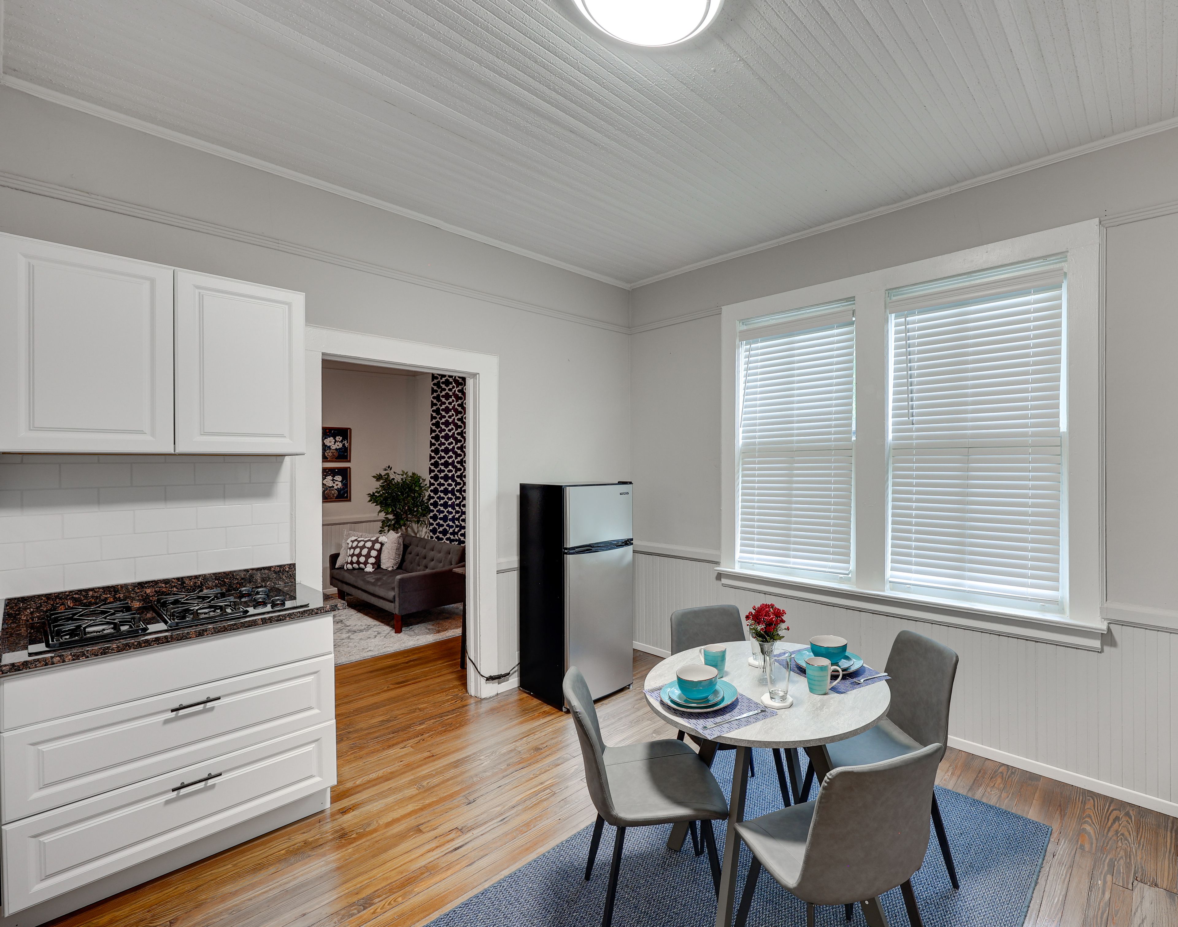 Une cuisine avec des meubles blancs, une cuisinière et un réfrigérateur. On y trouve également un coin repas avec une table, des chaises et de la vaisselle bleue.