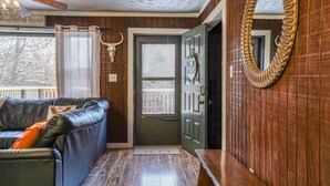 A room with a wooden floor, a black leather sofa with an orange pillow, a wooden bench, a door with a 'Hello' sign, and a mirror with a bull's head decoration. - Scenic Deep Creek Getaway Cabin-RICK'S RETREAT (Oakland)