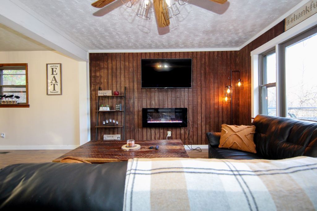 A cozy living room with a fireplace, a flat-screen TV, wooden panel walls, a leather couch, and a ceiling fan.