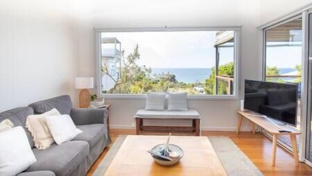 A modern living room with a grey sofa, a wooden coffee table, and a large window offering a view of the sea and buildings.