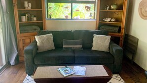 Un salon avec un canapé vert foncé, deux coussins décoratifs, une table basse en bois sur laquelle sont posés des magazines, et une fenêtre donnant sur un paysage verdoyant, avec une petite statue d’éléphant.