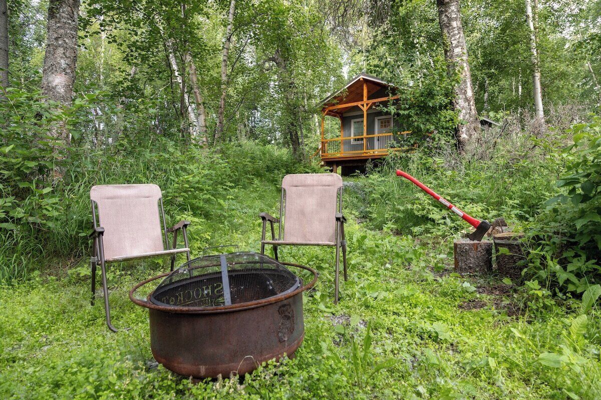 En enkel stuga omgiven av tät skog, med två stolar och en eldstad av metall i förgrunden.