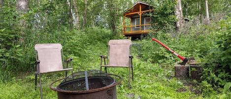 En enkel stuga omgiven av tät skog, med två stolar och en eldstad av metall i förgrunden.