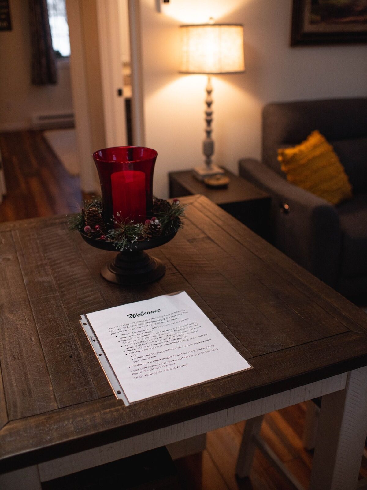 Une table en bois sur laquelle est posée une bougie rouge dans un joli photophore, un panneau d’accueil et une lampe en arrière-plan.