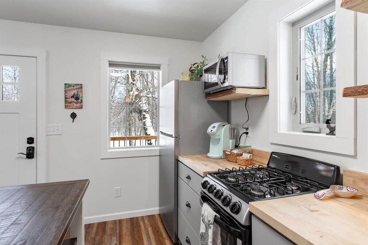 A modern kitchen with a gas stove, microwave, and refrigerator. There is a window with a view of trees, and a wooden table is visible in the foreground.