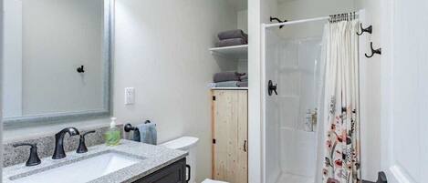 Un baño con lavamanos de granito, un espejo, inodoro, ducha con cortina floral y repisas con toallas.