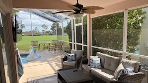 Ein Wintergarten mit Korbmöbeln, einem Deckenventilator und Blick auf einen Garten mit Pool und Terrassenmöbeln.