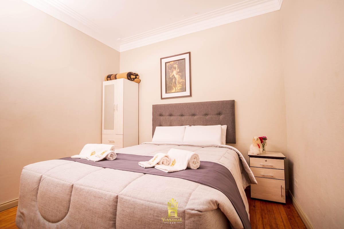 A neatly made bed with a grey headboard, a bedside table with a vase of flowers, and a framed picture on the wall.