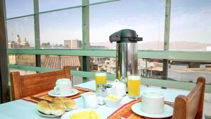 Ein Frühstückstisch mit Kaffeemaschine, Orangensaft und Teller mit Speisen, mit Blick auf Gebäude durch große Fenster.
