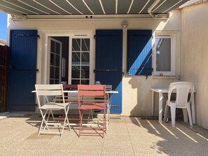 Ein überdachter Patio-Bereich mit Tisch und Stühlen, ein Fenster mit blauen Rollläden und eine Tür.