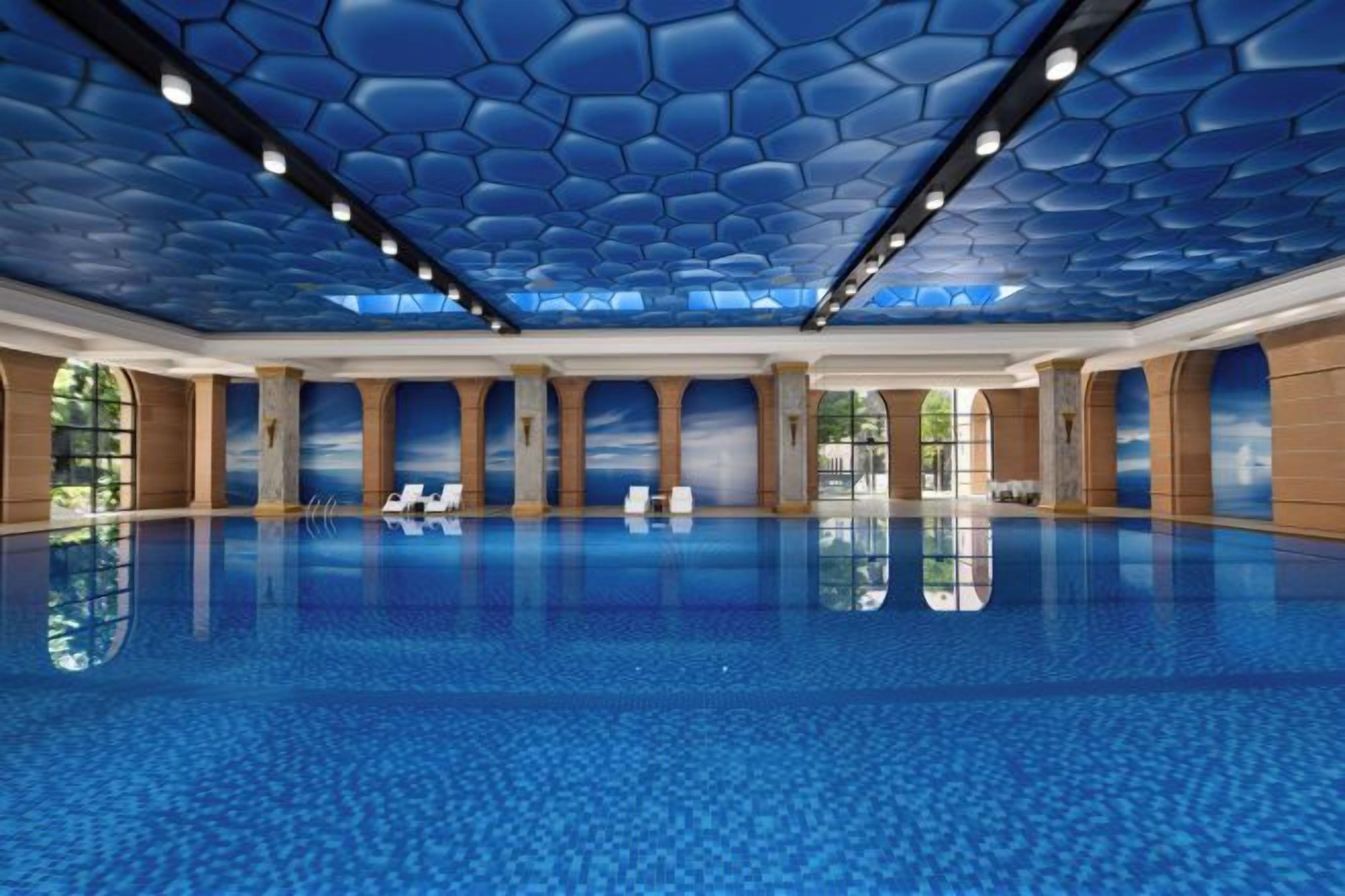 A large indoor swimming pool with a hexagonal ceiling design, clear glass walls, and a reflection of the pool on the floor.