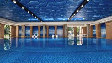 A large indoor swimming pool with a hexagonal ceiling design, clear glass walls, and a reflection of the pool on the floor.