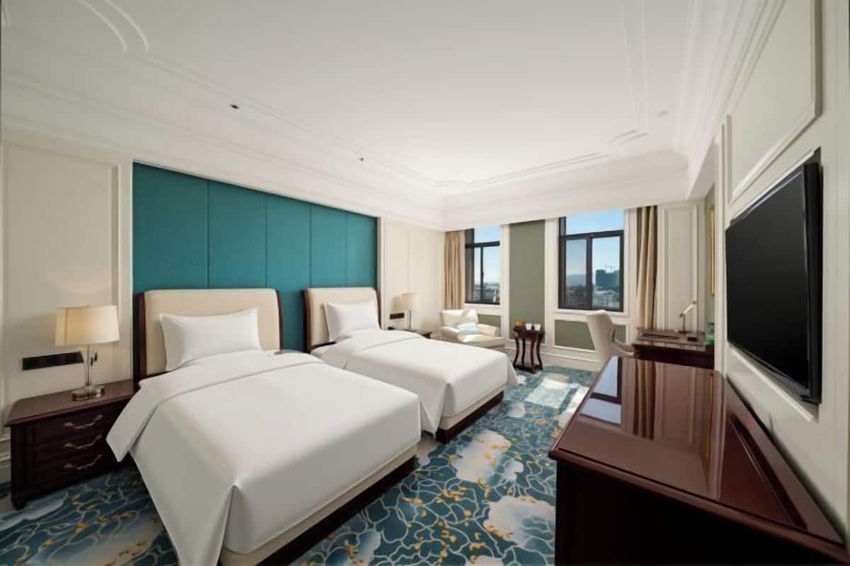 A hotel room with two beds, a desk, a TV, and a view of the city.