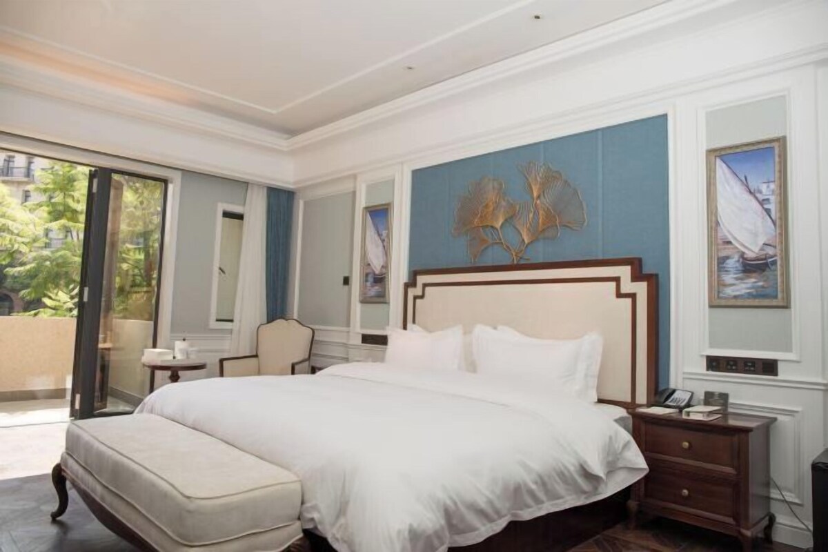 A hotel room with a large bed, a nightstand, a chair, and a view of greenery outside.