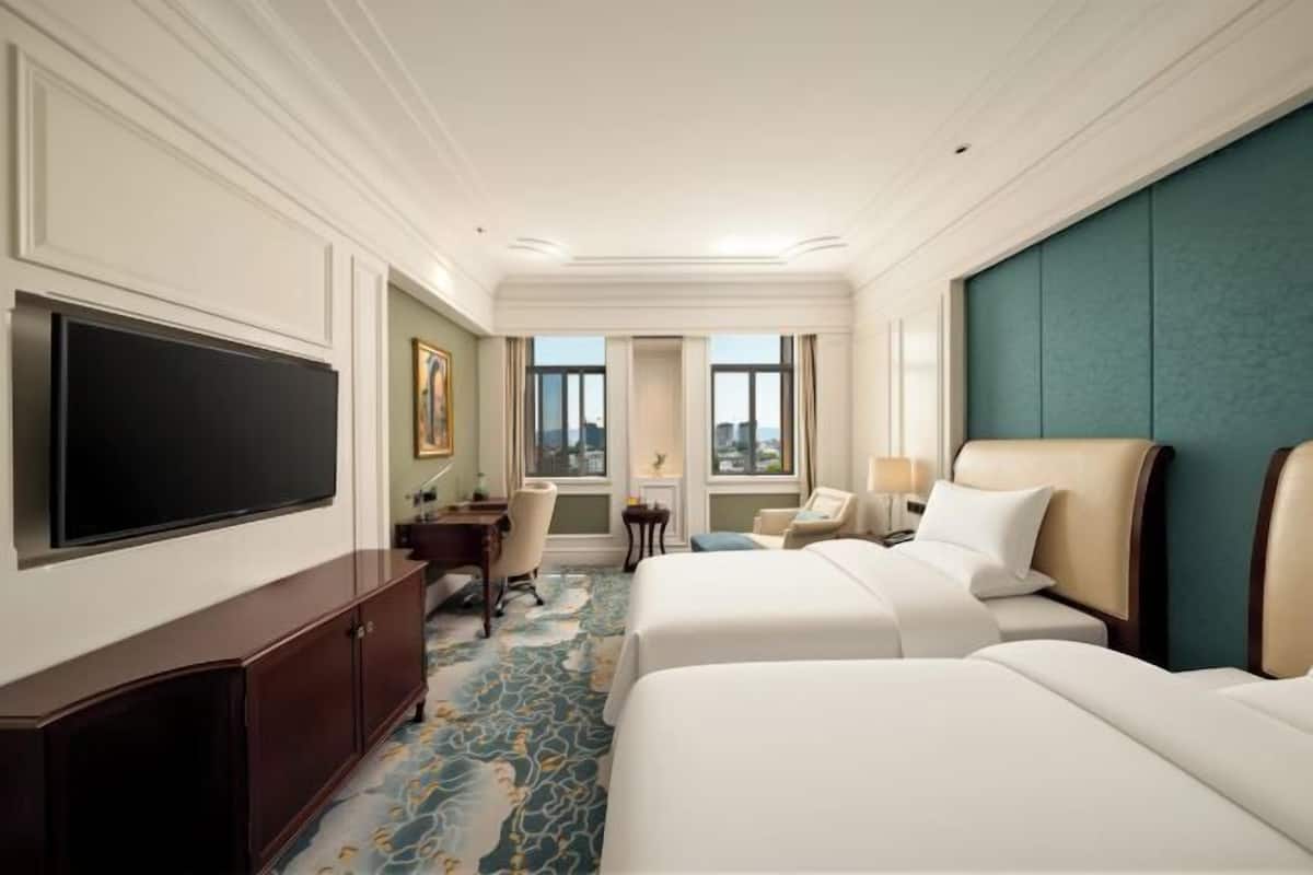 A hotel room with two beds, a desk, a TV, and a view of the city.
