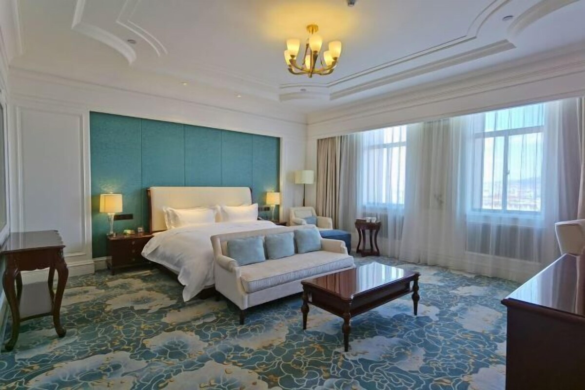 A hotel room with a large bed, a sofa, a coffee table, and a view of the city through large windows.