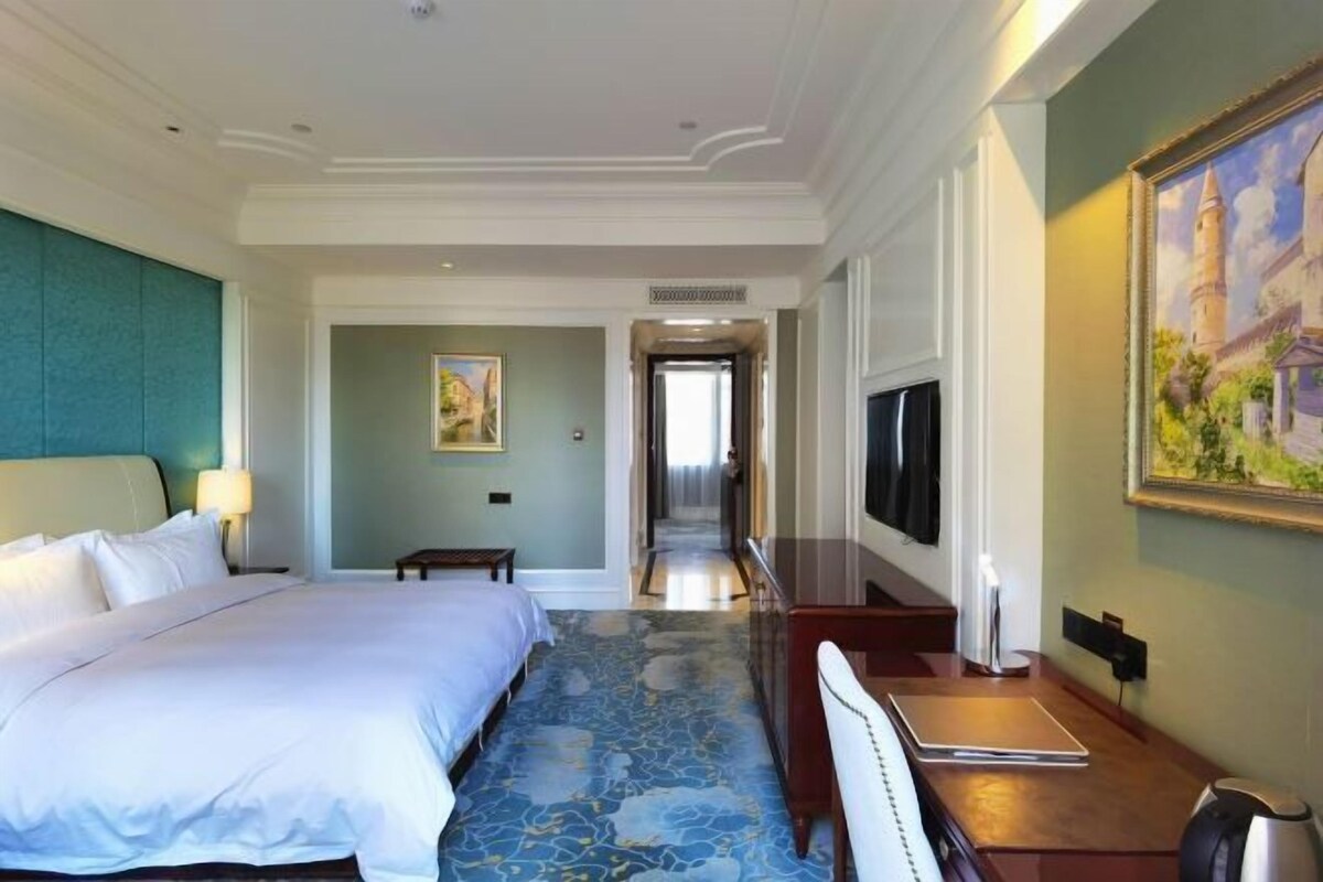 A hotel room with a large bed, a desk with a laptop, a TV, and a painting on the wall.