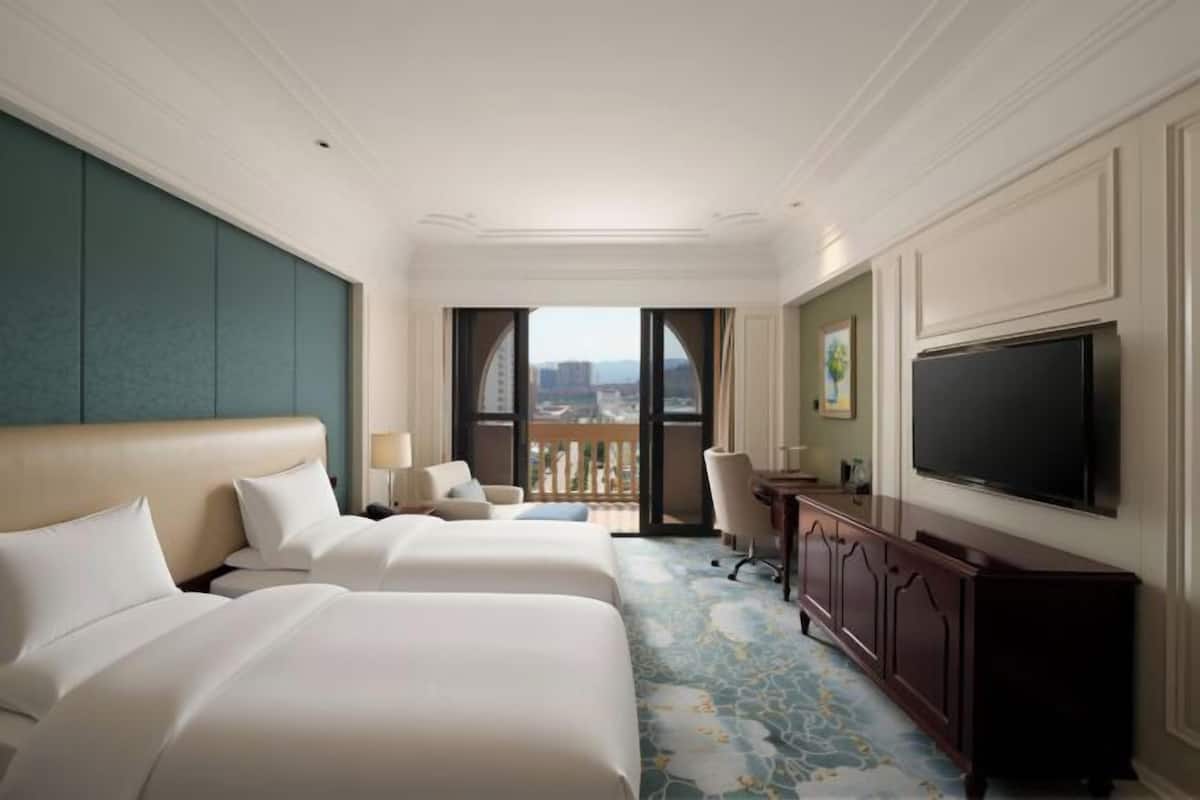 A hotel room with two beds, a TV, a desk, and a balcony with a city view.