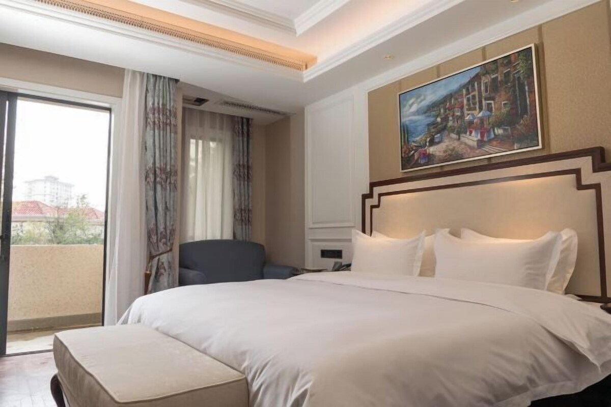 A hotel room with a large bed, a chair, a painting on the wall, and a view through a window.