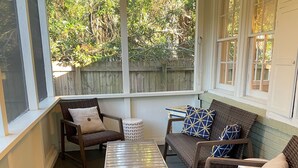 A covered patio with wicker furniture, a small table, and a view of a wooden fence and greenery.