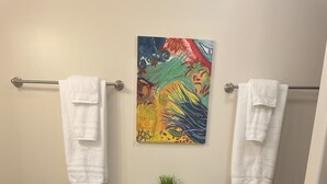 A bathroom with a toilet, a towel rack with two towels, a painting on the wall, and a small plant on the toilet tank.