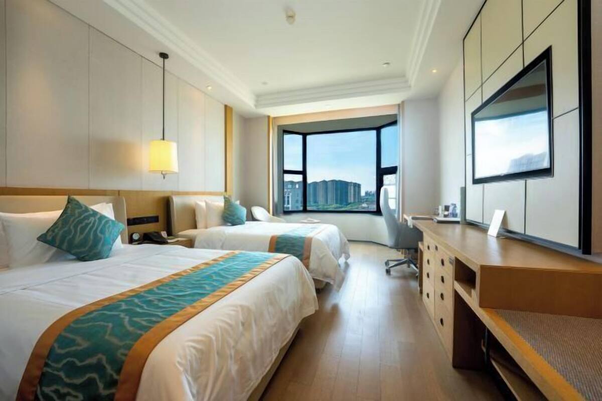 A hotel room with two beds, a desk, a chair, a TV, and a large window with a city view.